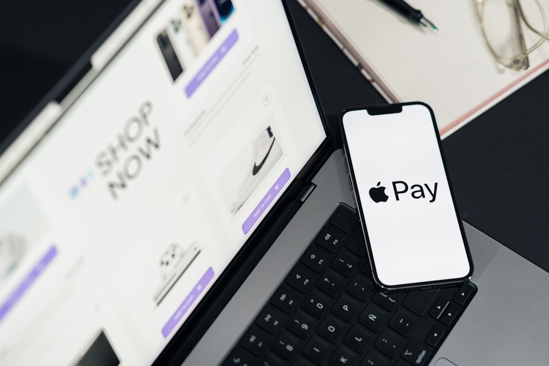 Apple Pay on phone screen. Top view of workspace. Table desk with laptop with ecommerce on screen, mobile, notepad